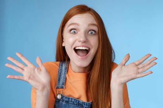 Amused speechless surprised astonished redhead girl hear exciting positive news smiling gladly wide eyes thrilled raised hand triumphing win lottery celebrating victory success, blue background.