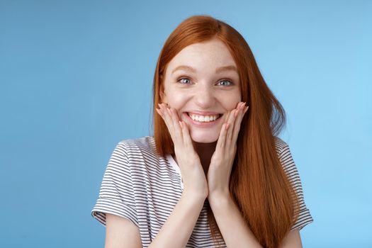 Excited happy cheerful redhead ginger girl smiling joyfully touch cheeks surprised receive amazing perfect b-day gift standing astonished triumphing rejoicing standing blue background. Copy space