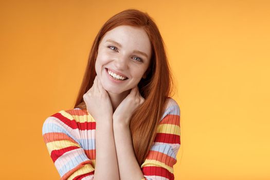 Silly enthusiastic attractive redhead blue-eyed girl tilting head touching neck flirty smiling enjoy perfect day feel happiness joy giggling coquettish flirting boyfriend, standing orange background.