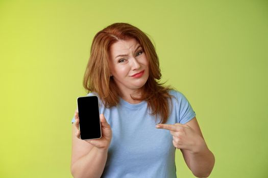 Seriously it awful. Displeased disappointed redhead mature female tilt head cringe grimacing reluctant pointing smartphone blank display index finger showing bad photograph share negative opinion.