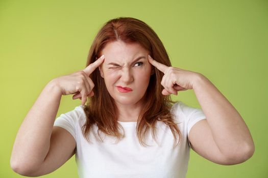 Difficult decision. Perplexed hesitant redhead middle-aged 50s woman trying solve puzzle look puzzled troubled smirk close eyes peek right touch temples thoughtful thinking intense green background.