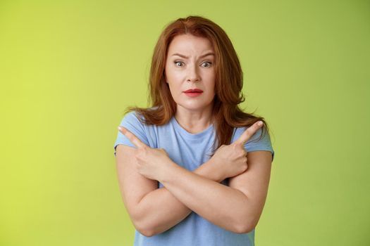Help me choose. Worried unsure doubtful cute redhead middle-aged silly woman asking advice cross arms body pointing sideways left right look concerned camera make difficult decision green background.