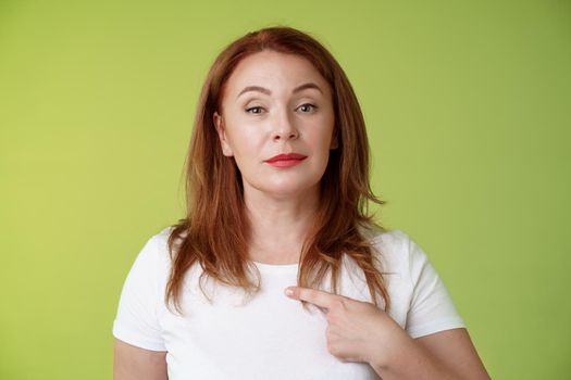 Proud mom pointing herself. Motivated confident redhead assertive middle-aged woman indicating chest volunteering bragging own abilities achievements look camera self-assured green background.