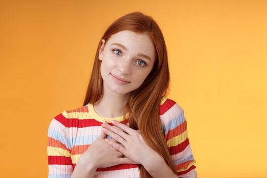 Lovely silly cute redhead young european girl tilting head look gently smiling tender camera press palms heart thankful receive nice gift gazing gratitude happiness, standing orange background.