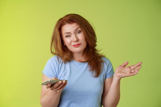 Well meh. Indifferent careless hesitant redhead middle-aged woman mature red female shrugging hold smartphone smirk bored uninterested hold hand aside apathetic attitude green background.