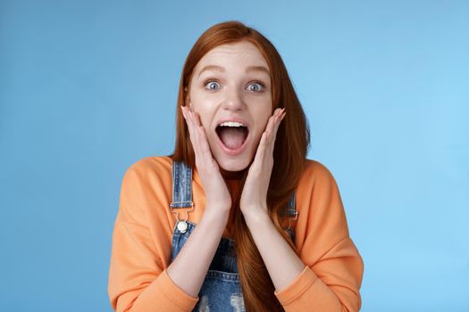 Lifestyle. Excited thrilled young emotional enthusiasitc ginger girl teenage college student yelling amused smiling broadly receive positive good news look surprised camera touch face astonished blue background.