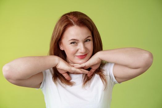 Aging, cosmetology, wellbeing concept. Happy self-assured redhead woman hold hands under jawline smiling showing facial blemished self-accepting wrinkles applying skincare product green background.