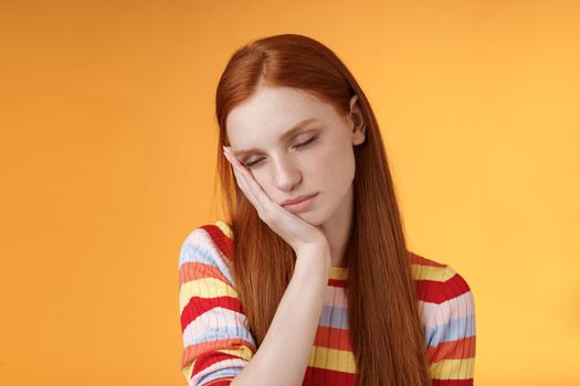 Tired cute redhead female student exhausted feel sleepy fall asleep standing leaning face palm close eyes working part-time night shift, daydreaming lacking energy wanna sleep bed orange background.