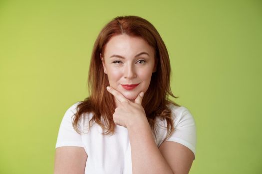 Hmm interesting choice. Intrigued cunning redhead middle-aged female hold hand face-line rub chin thoughtful smirking pleased raise eyebrow curious pondering alluring decision green background.