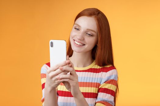 Cute feminine tender young redhead glamour girl holding smartphone taking pictures summer urban vibes female blogger shooting post online story standing happily orange background smiling.