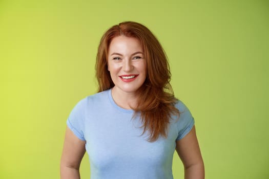 Cheerful kind happy redhead middle-aged mother look caring delighted smiling broadly gaze admiration joy stand blue t-shirt green background amused enthusiastic expression. Copy space