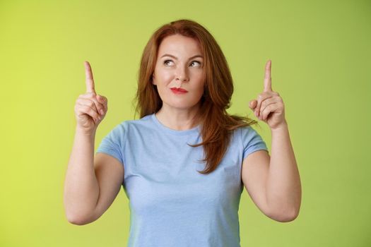 Curious thoughtful cute redhead female middle-aged ginger mother smirking thinking look aside pointing up index fingers deciding choice pondering choosing between variants green background.