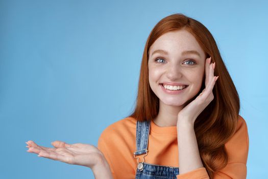 Cute tender chatismatic glad smiling redhead girl presenting awesome product show object palm hold hand raised blank copy space grinning impressed receive silly charming gift, blue background.