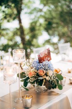 Bouquet in a vase on a table of pink and white roses and blue hydrangea, with the number of the table on the plate. Burning candles in high glass candlesticks.