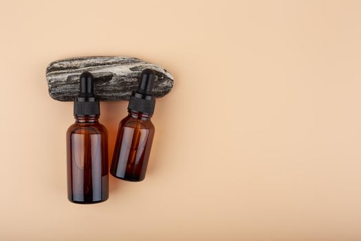 Two dark transparent glass bottles with skin serum or oil for manicure on black natural stone against bright beige background with copy space.