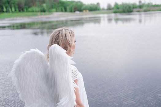 charming blonde with white angel wings standing by the water. beautiful woman in angel costume. goddess.