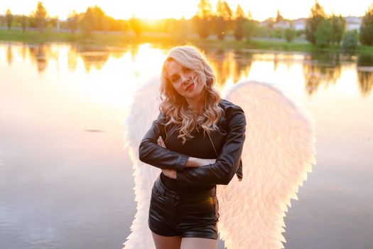 sexy blonde woman in black leather jacket and shorts with white angel wings. demon or angel in hell or heaven. sunset near lake.