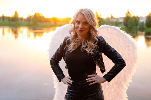 sexy blonde woman in black leather jacket and shorts with white angel wings. demon or angel in hell or heaven. sunset near lake.