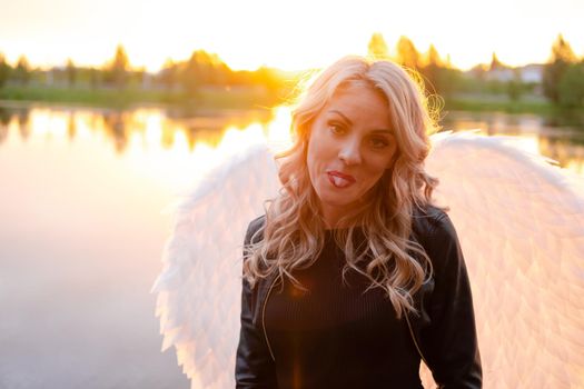 sexy blonde woman in black leather jacket and shorts with white angel wings. demon or angel in hell or heaven. sunset near lake.