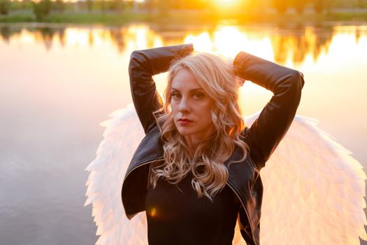 sexy blonde woman in black leather jacket and shorts with white angel wings. demon or angel in hell or heaven. sunset near lake.