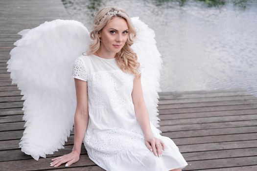 charming blonde with white angel wings standing by the water. beautiful woman in angel costume. goddess.