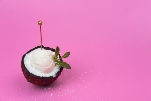 Vanilla ice cream balls in a fresh coconut half decorated with mint leaves with a spoon on a pink background.
