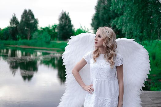 charming blonde with white angel wings standing by the water. beautiful woman in angel costume. goddess.