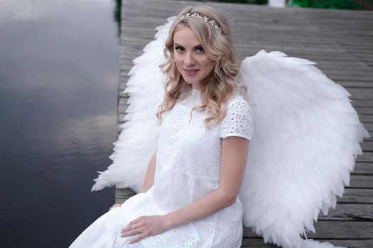 charming blonde with white angel wings standing by the water. beautiful woman in angel costume. goddess.