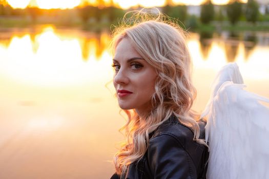 sexy blonde woman in black leather jacket and shorts with white angel wings. demon or angel in hell or heaven. sunset near lake.