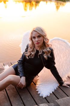 sexy blonde woman in black leather jacket and shorts with white angel wings. demon or angel in hell or heaven. sunset near lake.