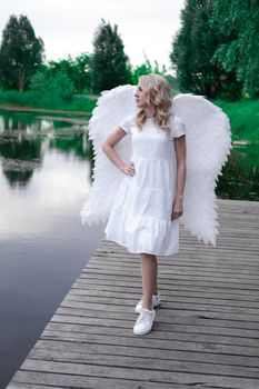 charming blonde with white angel wings standing by the water. beautiful woman in angel costume. goddess.