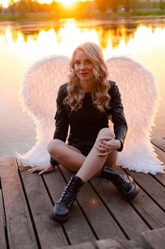sexy blonde woman in black leather jacket and shorts with white angel wings. demon or angel in hell or heaven. sunset near lake.
