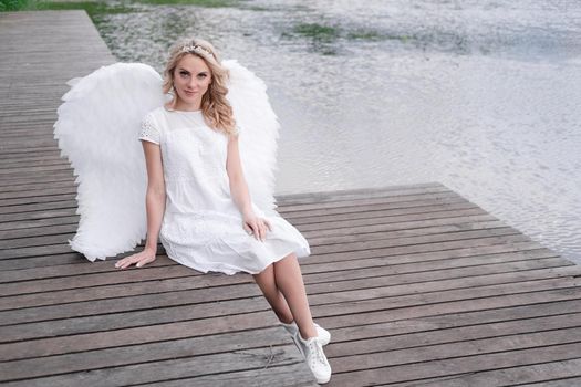 charming blonde with white angel wings standing by the water. beautiful woman in angel costume. goddess.