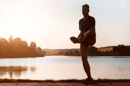 A young man trains outdoors against the backdrop of a beautiful lake at sunset, the athlete warms up, does exercises on the legs before running.
