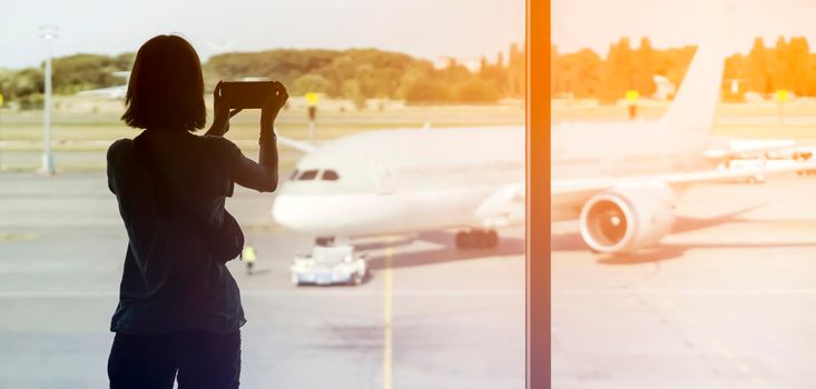 A young girl takes a photo on her phone before boarding the plane, the traveler goes on a trip, a silhouette at the airport by the window against the background of the aircraft.