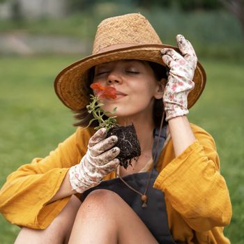 A young happy woman gardener in a straw hat and hands in gloves is holding a petunia flower in a peat pot, sniffing the flower and smiling before planting a seedling