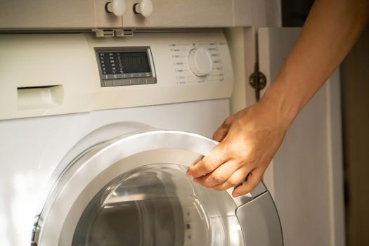 A male hand opens the door of a new washing machine in his modern, bright bathroom, close-up. The man washes clothes in the laundry.