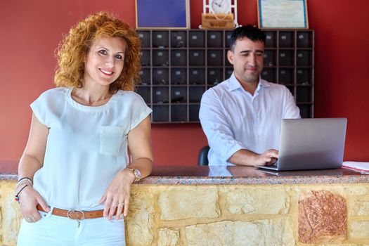 Small family hotel business, couple of hotel owners working at front desk. Smiling middle aged woman looking at camera and man working with laptop at reception, ownership, work, booking, accommodation