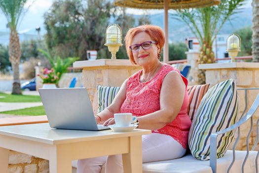 Portrait of elderly 70s woman with laptop sitting in outdoor cafe. Smiling senior female with glasses looking, reading, talking on video connection, resting. Technology, lifestyle, retirement age