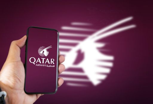 Doha, Qatar, July 2021: A hand holding a phone with the Qatar Airways airline app on the screen and the arabian oryx logo blurred on a dark cherry background.