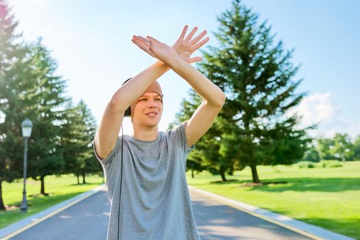 Portrait of cheerful hipster guy in knitted hat with headphones, dancing, hands up. Smiling teenager listening to music, summer in park. Hobbies, leisure, creativity, youth, lifestyle and technology