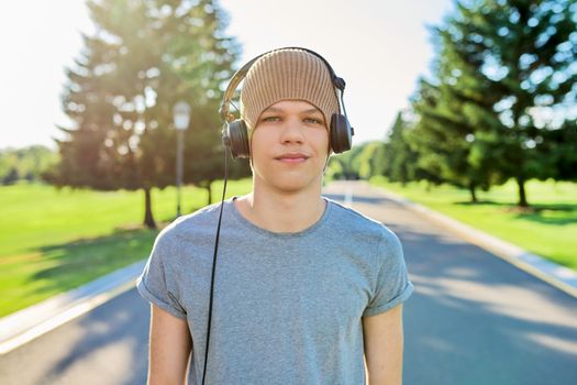 Portrait of teenage hipster in knitted hat with headphones looking in camera. Smiling guy listening to music, summer green nature in park. Education, leisure, adolescence, lifestyle and technology