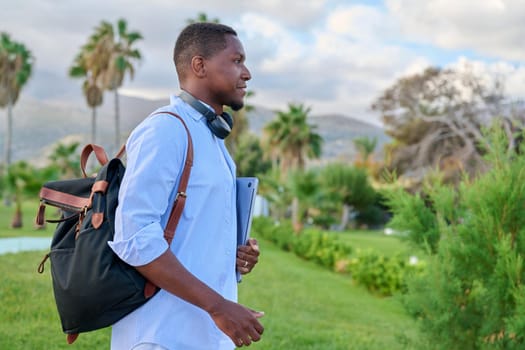 Outdoor portrait of young man with laptop headphones and backpack. African male adult student, creative freelancer, businessmen in tropical park. E-learning, online technologies, education, business