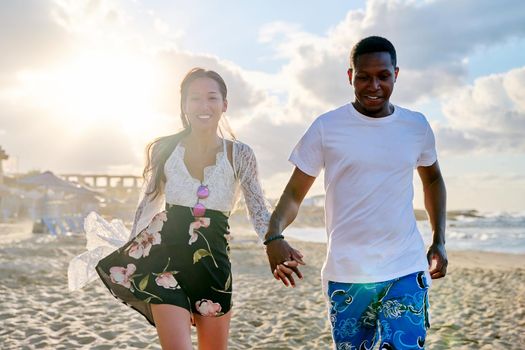 Beautiful young multiculturial couple running along beach together. Happy loving african man and asian woman holding hands. People, love, happiness, relationship, multiethnic family, vacation, tourism