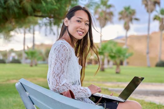 Young beautiful woman outdoors with laptop, tropical park background. Asian female sitting on bench, working, studying remotely. Freelance e-learning, online technologies, education, business concept
