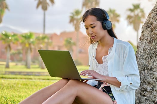 Young woman in headphones working on laptop outdoors. Asian female university student studying remotely, creative freelancer, business woman, blogger vlogger, work and study online, modern technology