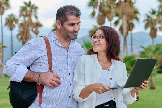 Middle-aged man and woman looking in laptop screen outdoors. Couple of business colleagues talking discussing smiling, tropical park nature background. Business, people, technology, mature age concept