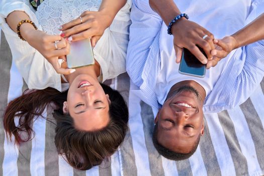 Top view smiling young couple looking at smartphone screens. Multiculturial couple of asian woman and african man outdoor vacationing, lying with phones in hand. Lifestyle, leisure, technology, people