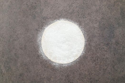 Thin homemade pizza dough with scattered wheat flour on dark concrete background.