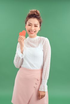Cheerful young Asian woman holding red heart paper isolated over green background. 